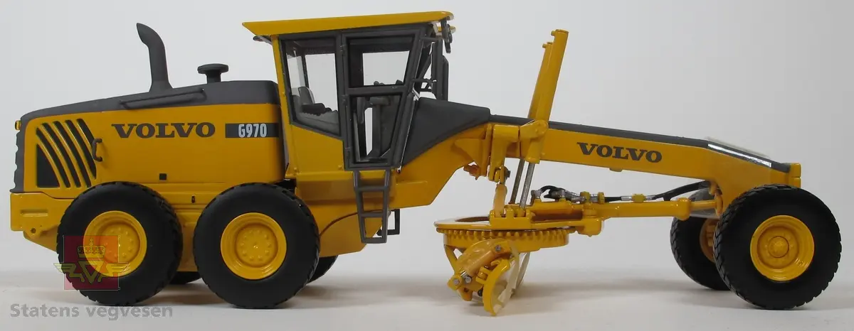 Volvo G970. Miniatyrmodell av veghøvel i eske. Esken har fargene svart, grå og burgunder samt påskriftene MOTORART collectible models og VOLVO. Den har også en skade i et av hjørnene der pappen har revnet. Modellen er laget hovedsakelig i metall med deler i plast og gummi. Fargen er gul med detaljer i svart og grått. Veghøvelen har tre akslinger, (seks hjul), og skjær. Merking fra produsenten under veghøvelen samt Volvo G970 merking utenpå. 
Skala 1:50.