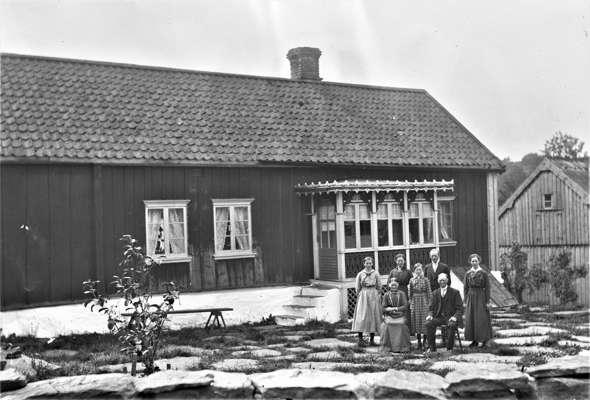 Sällstorp 6, gården kallas "Bernts". Anna-Kristina och Aron Johansson sitter vid manhusbyggnadens veranda. Bakom dem står från vänster barnen Emelia, Berta, Edit, Johan och Klara. Gårdsytan täcks av stora stenhallar och vid stengärdsgården står ett ungt träd. Verandan är dekorerad med intrikat snickarglädje, särskilt runt takfoten. I bakgrunden skymtar en omålad ekonomibyggnad.