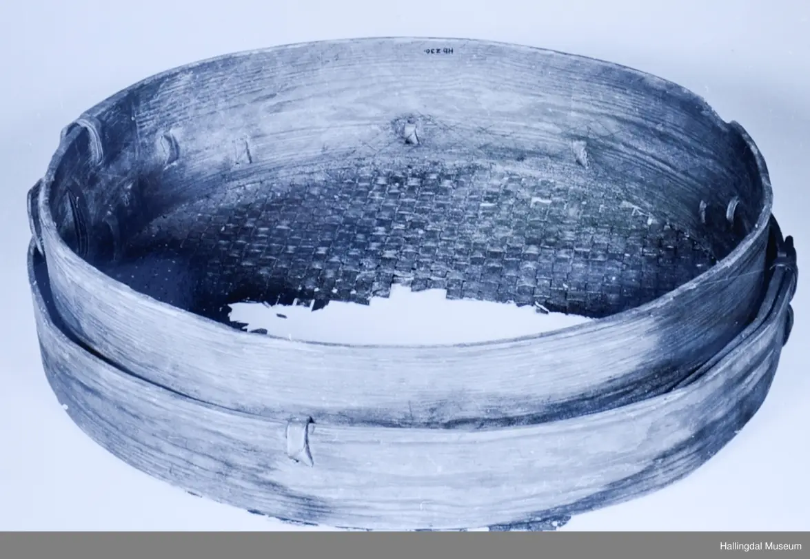 Tre, rundt. Siden dannet ett bøyd stykke, festet sammen med vidjer. Bunnen laget av smale, flettede spon, fastholdt ved utv. flatt, bredt trebånd.
R.B.S.