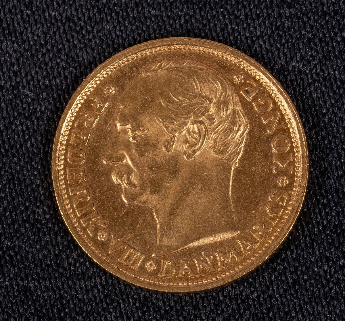 Danskt guldmynt, valör 10 kronor, från 1908. Ena sidan: Riksvapen, text: "1908 10 Kroner vbp". Andra sidan: Manshuvud i profil, text: "Frederik VII Danmarks Konge".