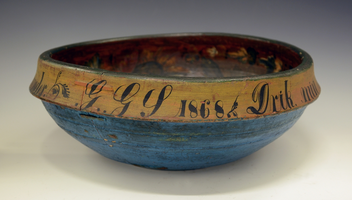 Rosemalt ølbolle. Fra protokollen: Ølbolle, svarvet med sterkt utstaaende ribbe, hvorfra en kant böier ind. Nyere overmaling, utenpaa malt lyseblaa med minjerød kant, hvorpaa indskriften: "Drik min Ven. Jeg dig det Under. Pust imelem og Giv dig stunder. G.G.S. 1868". Indeni er paa gulbrun bund  malt svære roser i c-oplæg og nærmest i Luraasstil; farver: rødbrunt, grønt, blaat, hvitt, gult; sort skrafering. Bollen er kinget.