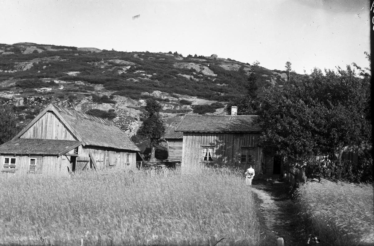 Grimmared, Låbäck. Gårdsbebyggelsen sedd på avstånd, alla byggnaderna är omålade. Utanför stugans farstukvist står husbonden och makan sitter bredvid honom med ett litet barn i knät. Åkrarna går nära in på husen och bakom gården reser sig ett berg.