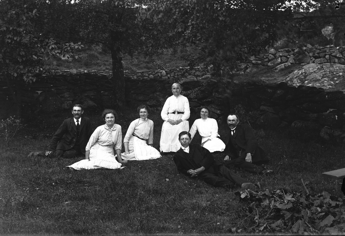 Kärskens i Grimmared; fr v. Axel, Tilda, Hulda, Josefin A på en stol, Julia, Birger och framför dem ligger gårdens dräng. De är samlade i trädgården där en hög stengärdsgård går upp längs slänten bakom dem. En hög med avklippta lönnkvistar ligger nere i högra hörnet vid en bänk.
(Se även bilderna GEA029, GEA112 och GEA115)
