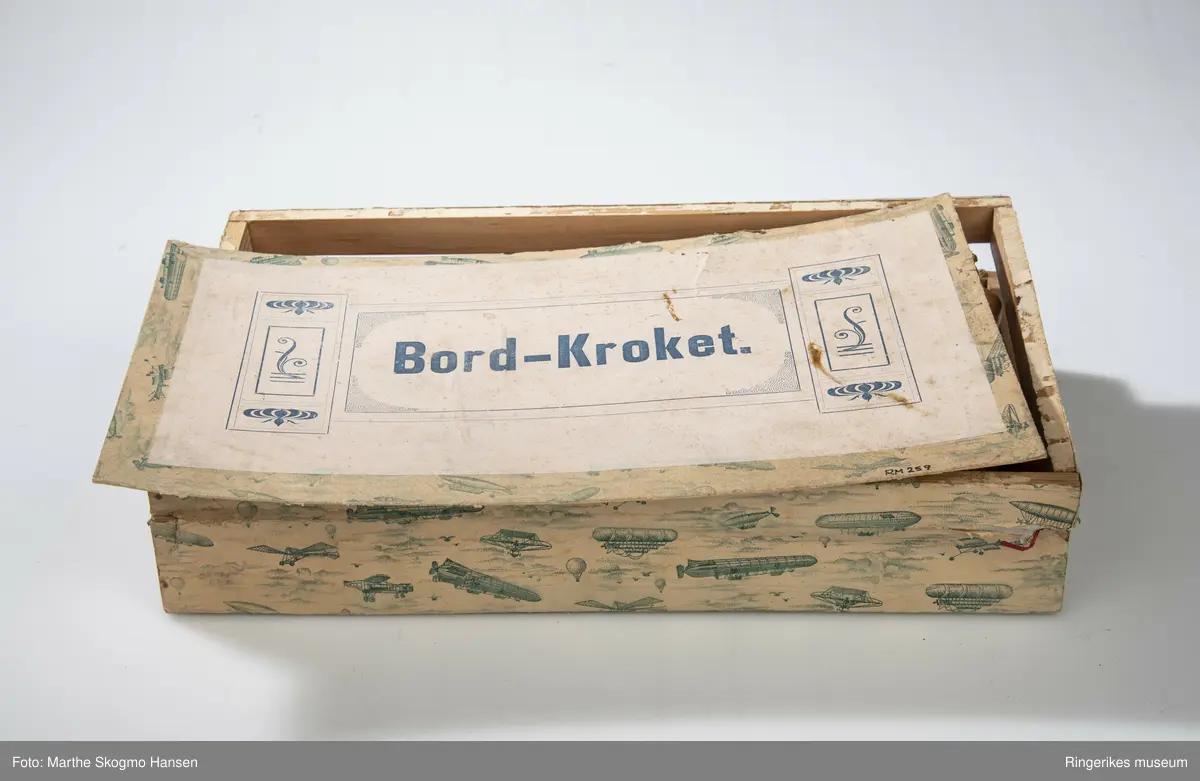 Treeske med bordkrocket. 8 køller og kuler i lakkert tre merket med fargene blå, svart, hvit, rød, gul, grønn, lilla og brun og 2 målpinner. 10 bøyler av ståltråd med røde metallføtter.
