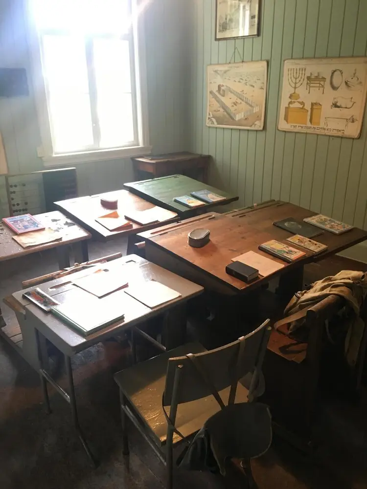 Skinnarbøl skolemuseum. Fotografi tatt inne i klasserommet. Veggen er lysegrønn, på den henger skoleplansjer. Fire gamle skolepulter.