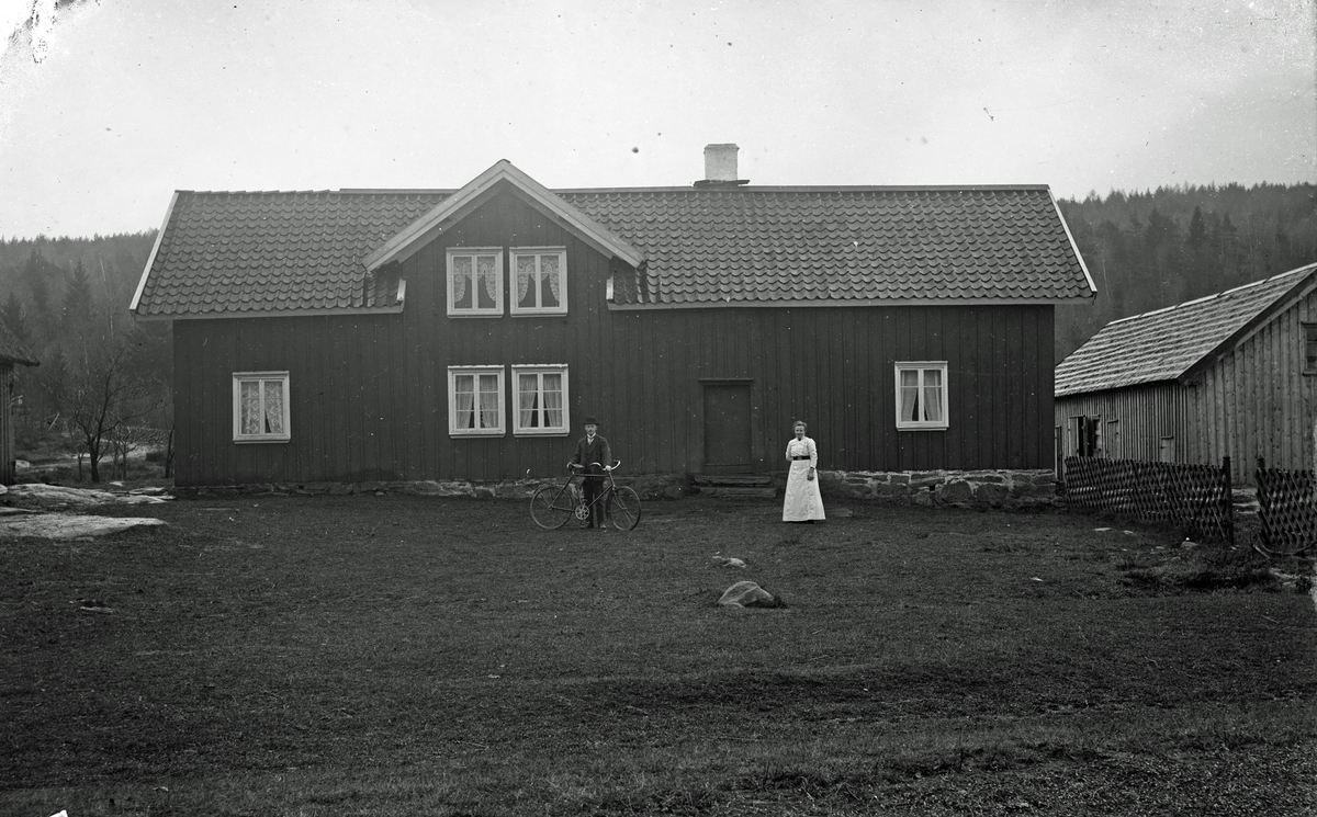 En man i kubb står med en cykel på ett gårdstun tillsammans med en kvinna. Manbyggnaden är långsträckt och målad, med en asymmetriskt placerad frontespis dit fönstren är koncentrerade. Från husgaveln till höger i bild går ett trästaket med korslagda spjälor och på utsidan av det ligger en ekonomibyggnad. Även till vänster skymtar ett hus och byvägen som slingrar sig upp mot en granklädd höjd.