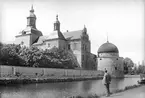 Visst är den imponerande, vasaborgen i Vadstena. Denna sommardag i början av förra seklet var amatörfotografen Carl Sundström och gissningsvis åtminstone en bekant på besök för att beskåda slottet och rimligtvis annat som den pitoreska staden kunde erbjuda dem. De hade inte åkt långt. Sundström var vid tiden handlare i Viby så sannolikt hade de tagit tåget från Mantorp. Slottet de besåg hade på beslut av Gustaf Vasa uppförts som försvarsanläggning i mitten av 1500-talet, men hade i mer fredlig tid fått skilda funktioner, så varierande såsom husrum för damastväveri som sädesmagasin. Från år 1899 gav slottet plats för Landsarkivet.