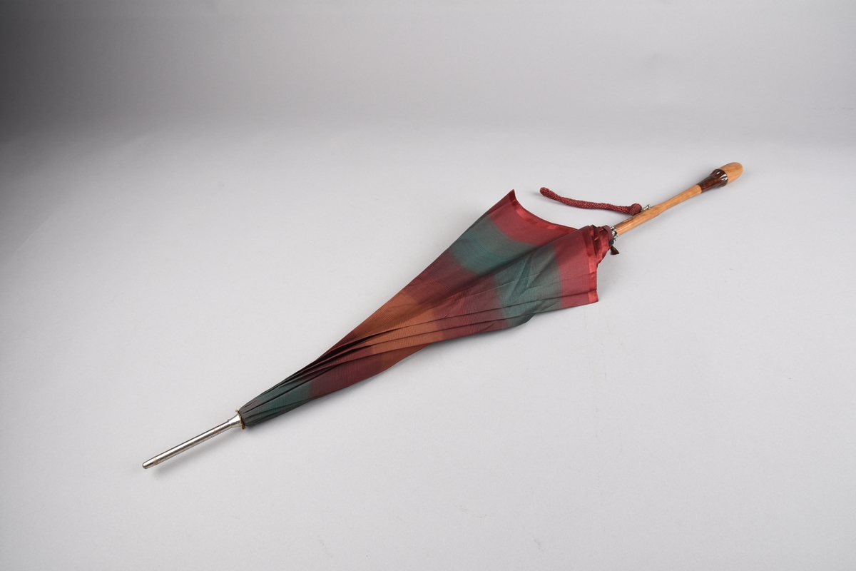 Paraply med 10 spilar og stong i metall. Handtak i ljost tre med ei mørkare tresort som dekorelement. Hempe i tekstil for å ha handa i, er festa på handtaket i ei metallring. Stripete duk i kunstfiber i fargane plommeraud, grøn, oransje og raud. Hempe for å lukka paraplyen att.