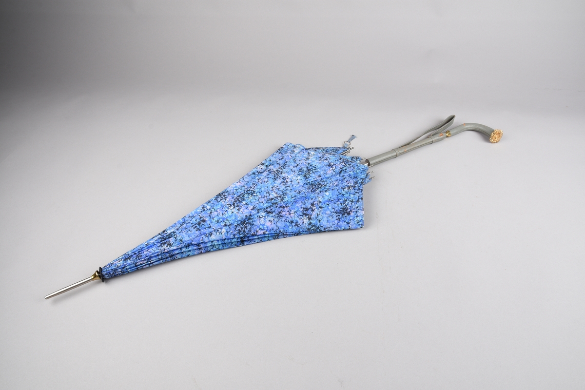 Paraply med 10 spiler og stang i metall.Handtak i skai med dekor i plast, hempe for å ha handa i er festa på handtaket. Duk i kunstfiber i fargane lilla, blå, svart og lys blå, spetta.