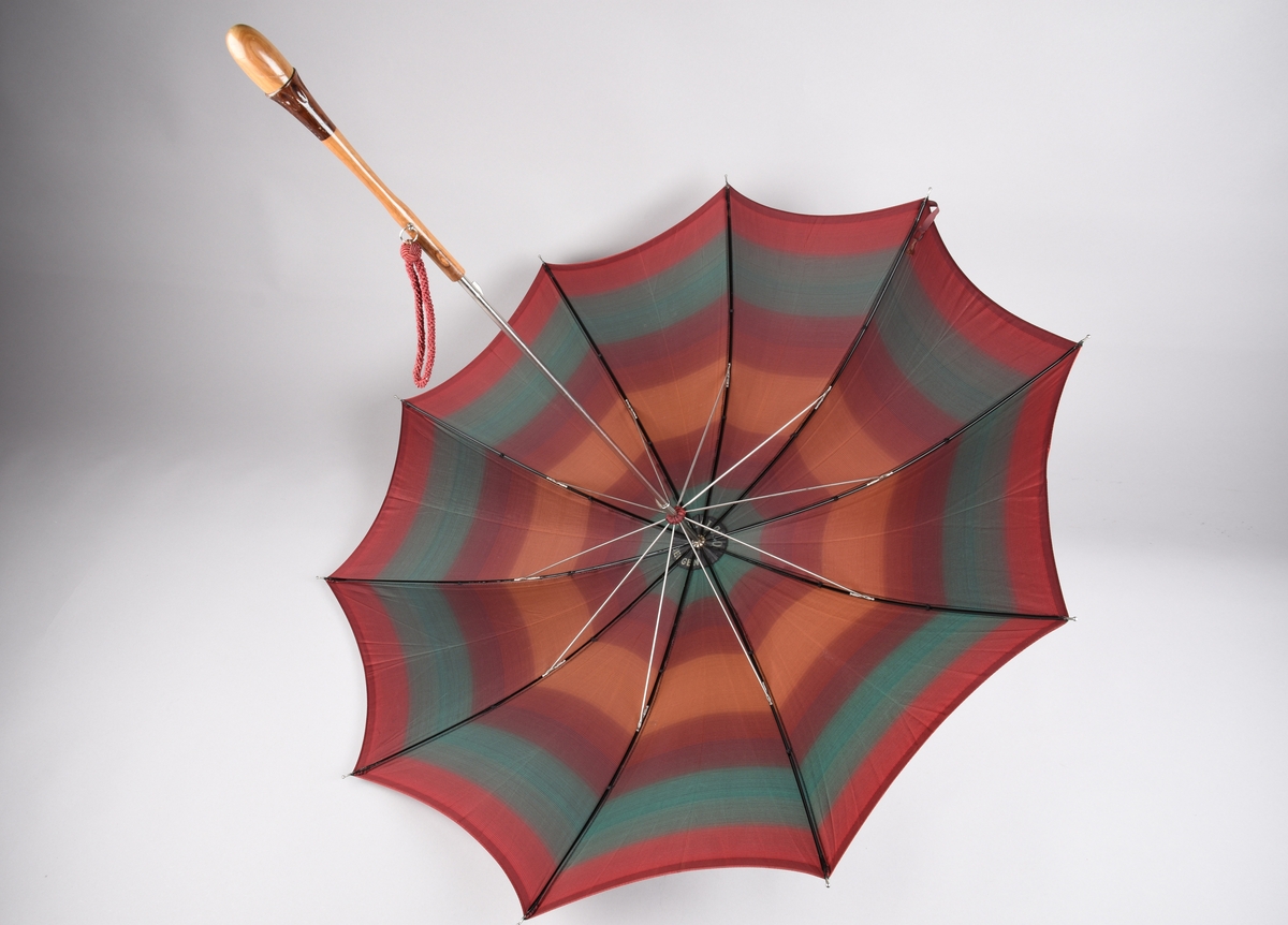 Paraply med 10 spilar og stong i metall. Handtak i ljost tre med ei mørkare tresort som dekorelement. Hempe i tekstil for å ha handa i, er festa på handtaket i ei metallring. Stripete duk i kunstfiber i fargane plommeraud, grøn, oransje og raud. Hempe for å lukka paraplyen att.