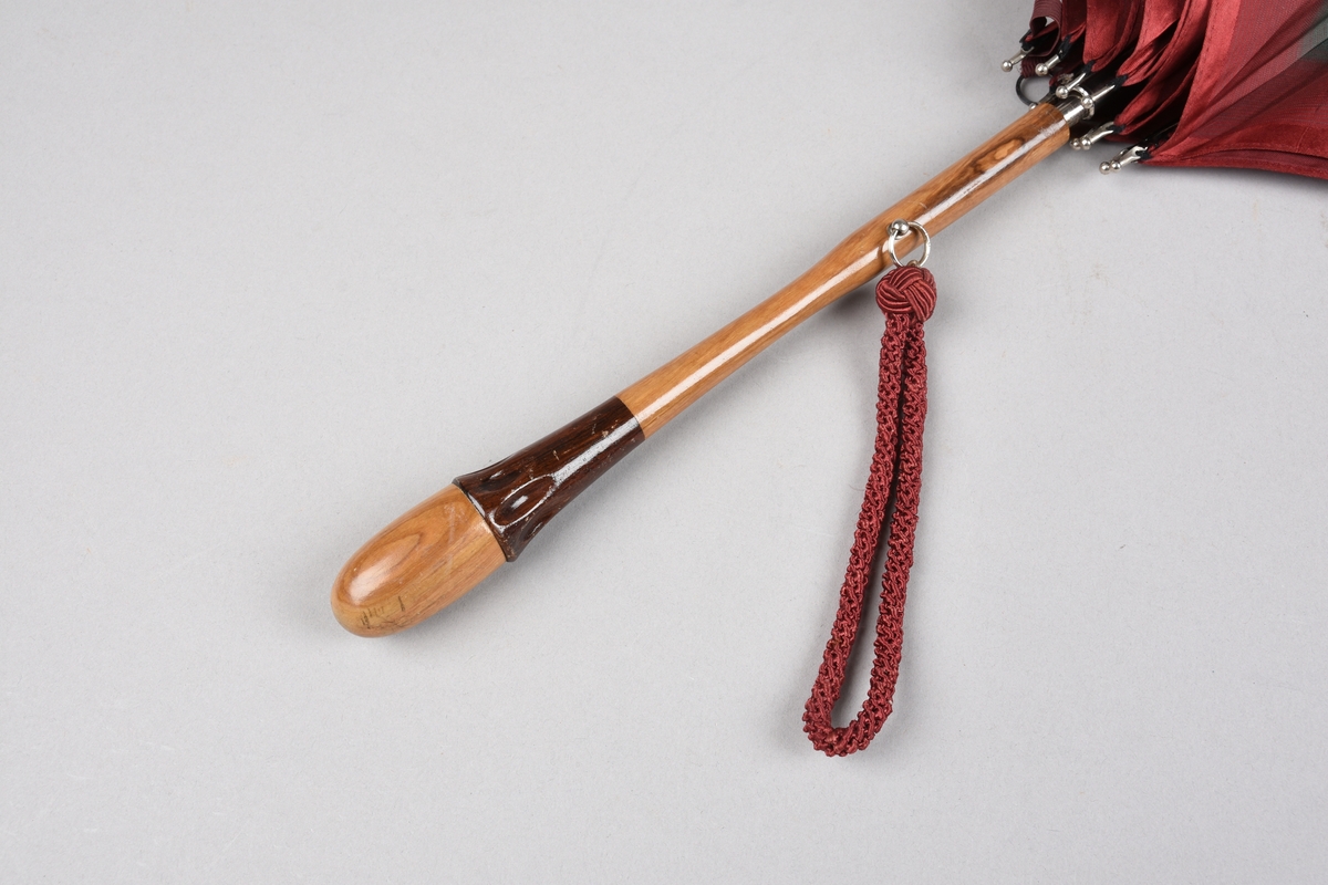 Paraply med 10 spilar og stong i metall. Handtak i ljost tre med ei mørkare tresort som dekorelement. Hempe i tekstil for å ha handa i, er festa på handtaket i ei metallring. Stripete duk i kunstfiber i fargane plommeraud, grøn, oransje og raud. Hempe for å lukka paraplyen att.