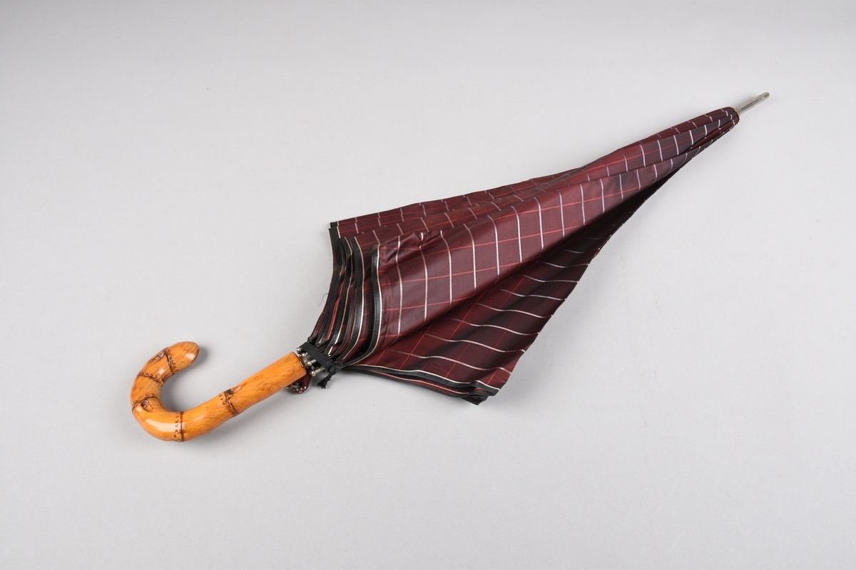 Paraply med 10 metallspilar. Stong i metall. Trekt med stoff i kunstfiber, rutemønster. Handtak i tre, imitert bambus, med hempe i tekstil til å ta inn på handa. Hempe for lukking av paraplyen.