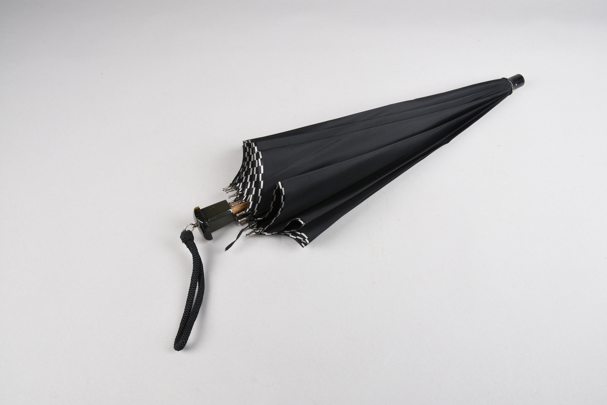 Paraply med 16 metallspilar. Stong i tre. Trekt med stoff i kunstfiber, ensfarga svart med dekor i sølv/svart rutemønster. Handtak i plast med hempe i tekstil til å ta inn på handa. Hempe for lukking av paraplyen.