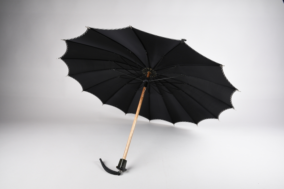 Paraply med 16 metallspilar. Stong i tre. Trekt med stoff i kunstfiber, ensfarga svart med dekor i sølv/svart rutemønster. Handtak i plast med hempe i tekstil til å ta inn på handa. Hempe for lukking av paraplyen.