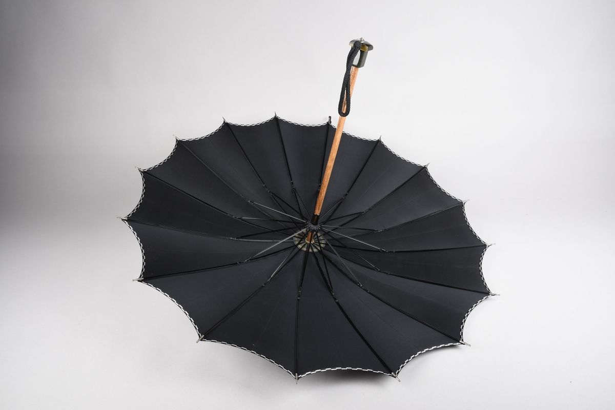 Paraply med 16 metallspilar. Stong i tre. Trekt med stoff i kunstfiber, ensfarga svart med dekor i sølv/svart rutemønster. Handtak i plast med hempe i tekstil til å ta inn på handa. Hempe for lukking av paraplyen.