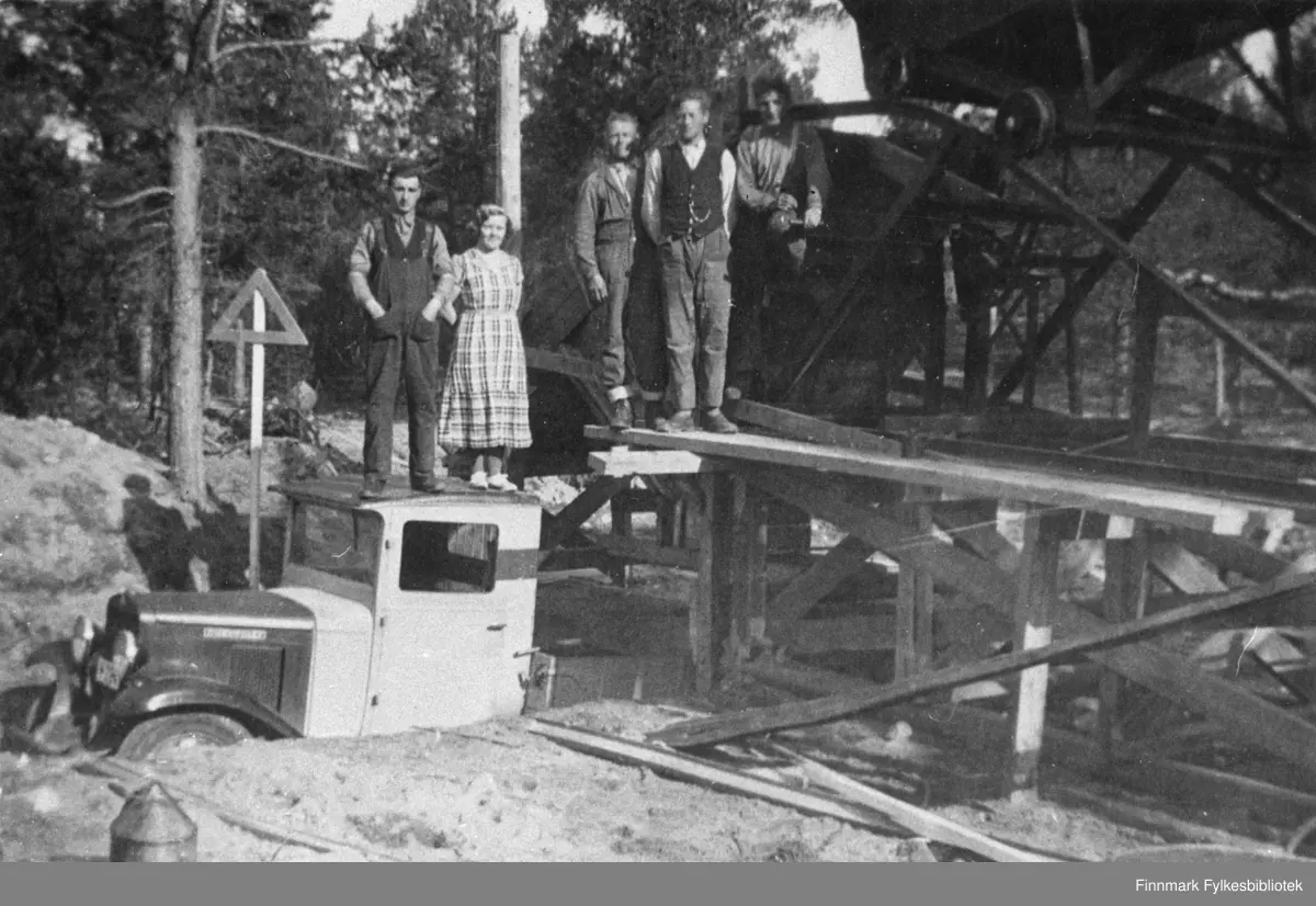 Grussortering i Nattvann i Karasjok i 1937. Gruslasteapparat fra Ankerløkken Mek. Verksted. Dette var tungvint i bruk. To personer står på taket til en lastebil (Chevrolet 1929 eller 1930 modell) og tre står på gruslasteapparatet. Fra venstre: Edvard Ratama, Margreta Mikkelsen, Otto Abrahamsen, Karl Grøtte og Anders Nymo.