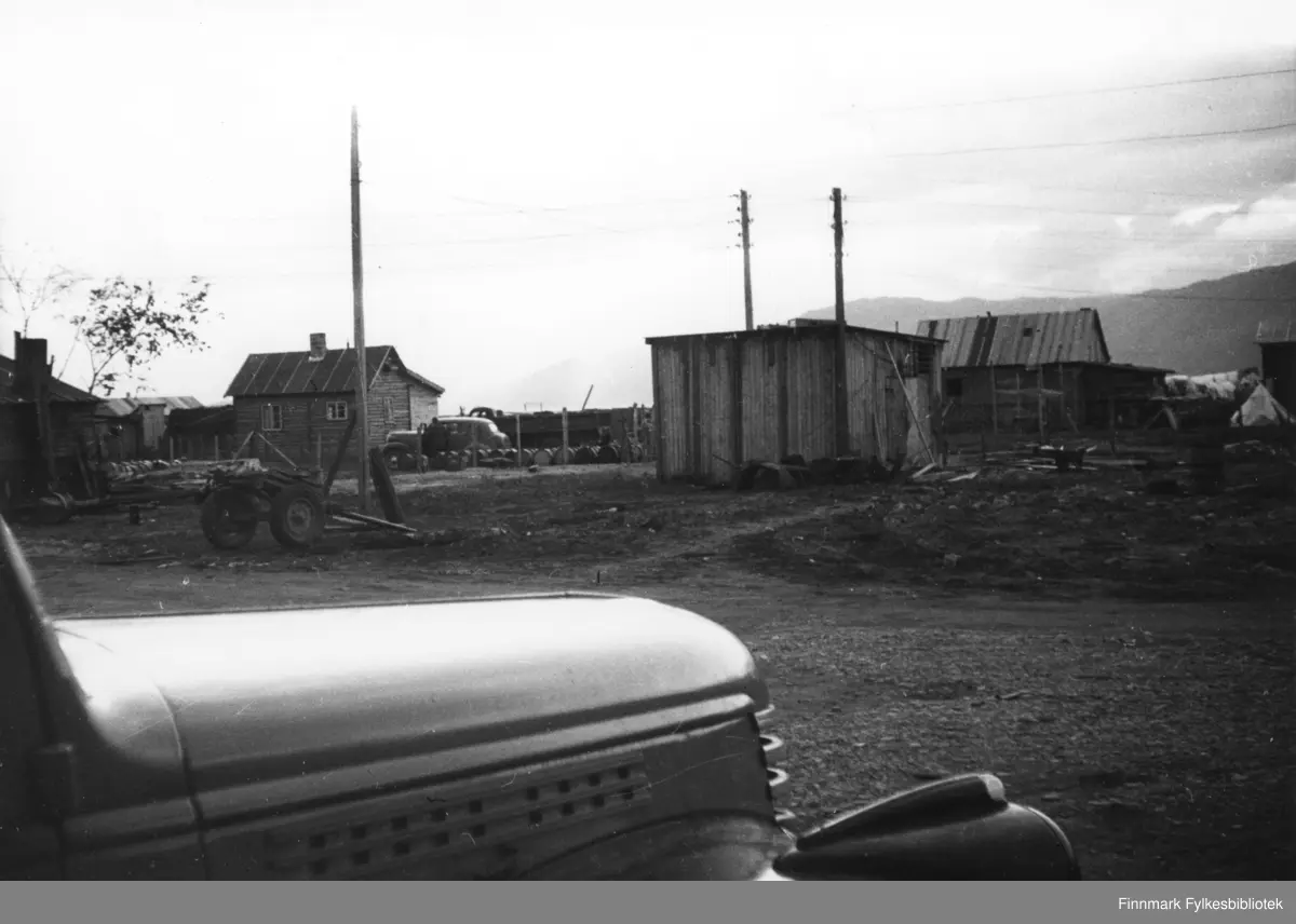 Fronten på en lastebil, en Chevrolet 1946-47 stikker inn i bildets forkant. I bakgrunnen ser man bebyggelse og en kjerre med hjul, flere telefonstolper rager opp i siluett mot himmelen. 
