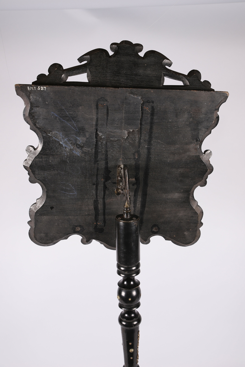 Notestativ i sortmalt tre. Notestol og fot kan settes sammen, foten deler seg i tre nederst. Rike profileringer på fot og stol, messingbeslag som er hamret i naturalistiske og stiliserte ornamenter. Stolen er på forsiden trukket med rustrødt, stoppet sikestoff. Denne kan reguleres på baksiden. Her kan men se merker etter to lister som sannsynligvis er det gamle festet mellom stol og fot.