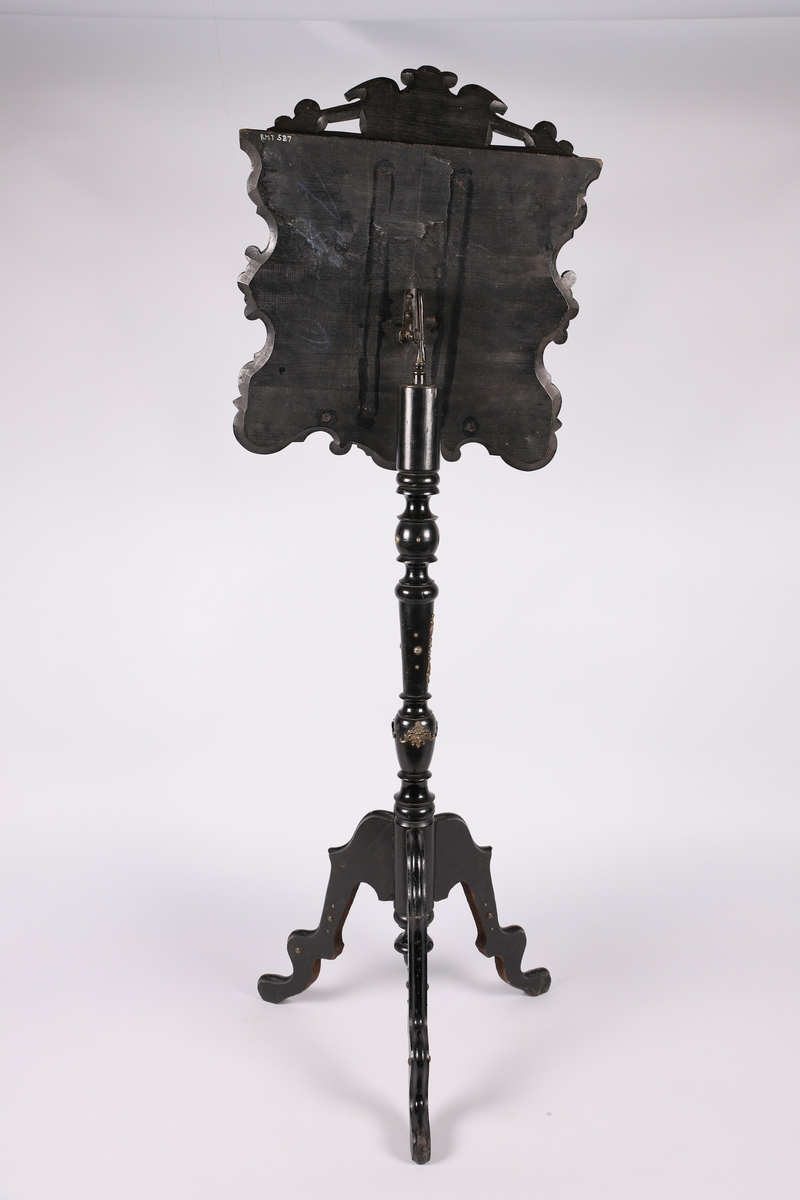 Notestativ i sortmalt tre. Notestol og fot kan settes sammen, foten deler seg i tre nederst. Rike profileringer på fot og stol, messingbeslag som er hamret i naturalistiske og stiliserte ornamenter. Stolen er på forsiden trukket med rustrødt, stoppet sikestoff. Denne kan reguleres på baksiden. Her kan men se merker etter to lister som sannsynligvis er det gamle festet mellom stol og fot.