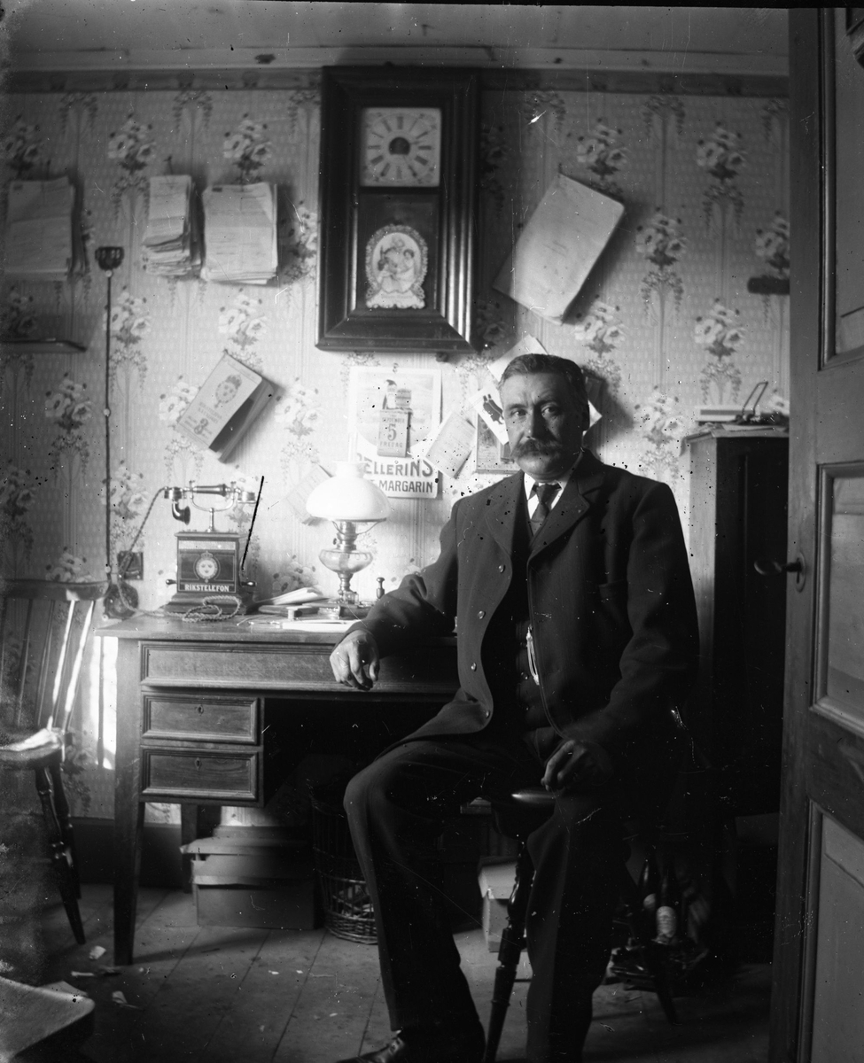 En man sitter vid ett skrivbord där det står en Rikstelefon. Ovanför den hänger en telefonkatalog och buntar med olika dokument. Almanackan med reklam för Pellerins margarin har inte många blad kvar, så troligen är det fredag den 5:e december och då kan året vara antingen 1913 eller 1919.