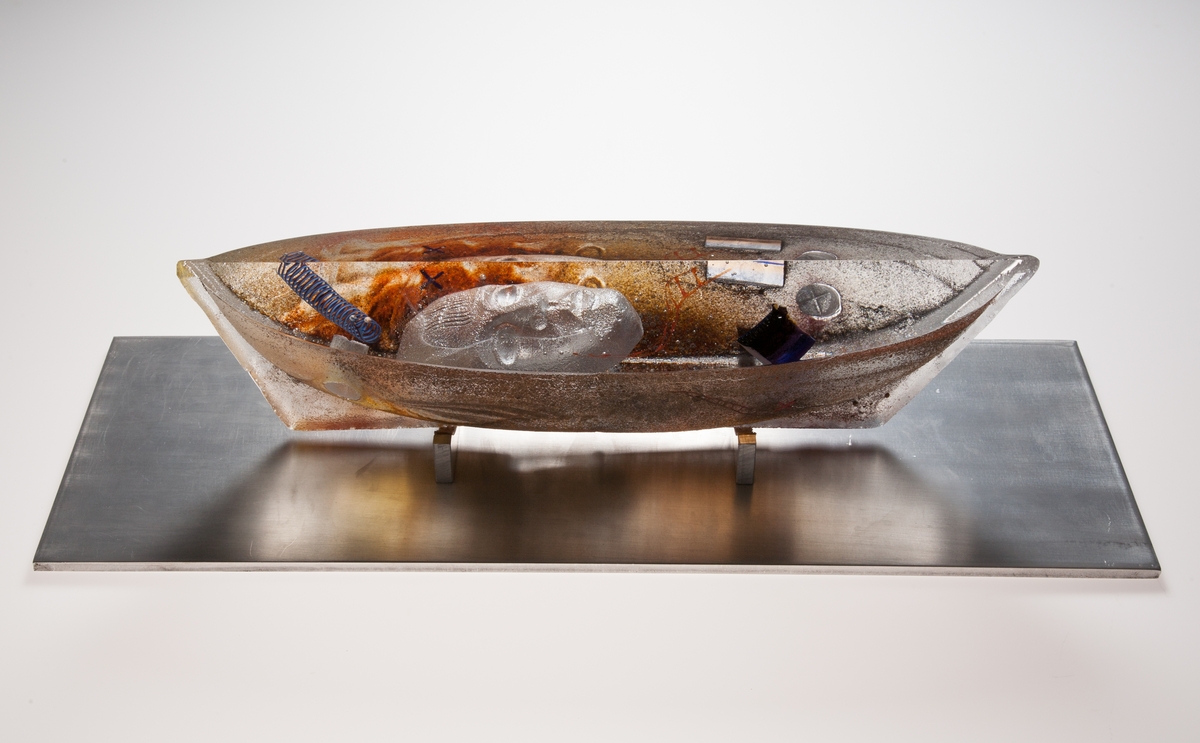 Glasbåt "Celestial Journey II" med tillhörande metallplatta.
Gjuten, sandig struktur på båtens yttre, ansikte dominerar båtens insida.
Brunaktig färg.