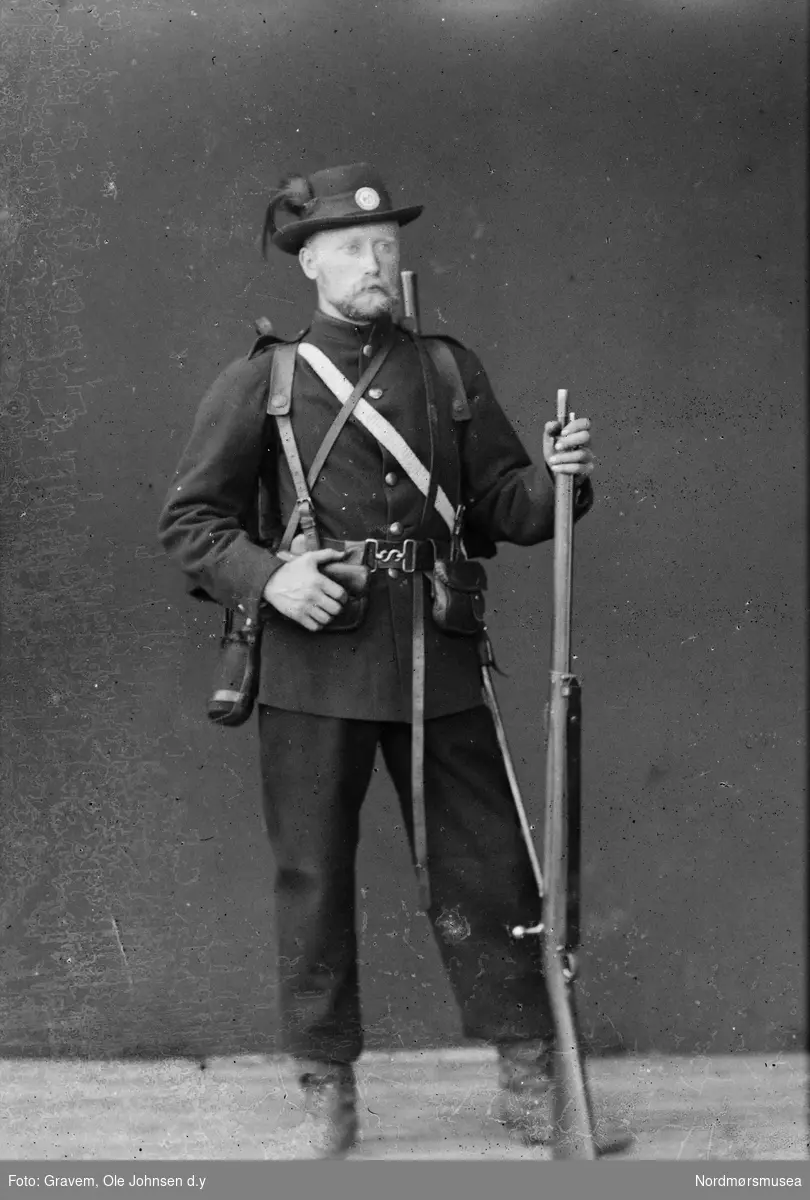 Portrett i helfigur av mann med uniform hatt og gevær. Dette er en infanterist (musketer) i uniform som reglementert fra 1877 til 1888.