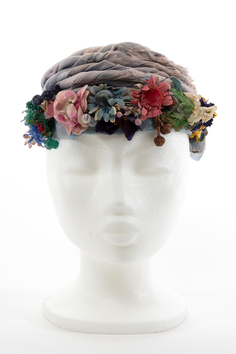 Lav, liten hatt uten brem, av lyseblå og dueblå silkechiffon. Pullen er lav og avrundet, konstruert av et grovt og mykt, hvitt nettstoff, dekket av flere lag lyseblå og tåkeblå chiffon, det ytterste laget dueblått. Mens toppen er glatt, er pullens skrå sider garnert med 5-6 snodde remser av dueblå og lyseblå chiffon. Pullens nederkant har en litt avstivet "linning", også den er trukket glatt med ciffon og nestet til resten av pullen for hånd. Nederkanten er prydet med en tett, polykrom krans av varierte blomster og blader av silke, og bær av hardt, vokset materiale. Innvendig fôret med kremfarget silke, festet for hånd, men figursydd med maskin. Svart, rund hattestrikk festet i begge sider.