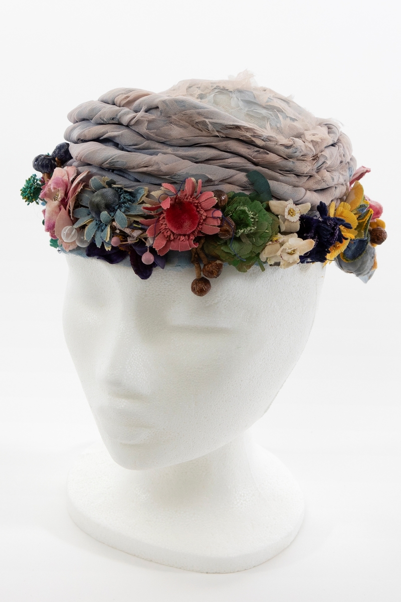 Lav, liten hatt uten brem, av lyseblå og dueblå silkechiffon. Pullen er lav og avrundet, konstruert av et grovt og mykt, hvitt nettstoff, dekket av flere lag lyseblå og tåkeblå chiffon, det ytterste laget dueblått. Mens toppen er glatt, er pullens skrå sider garnert med 5-6 snodde remser av dueblå og lyseblå chiffon. Pullens nederkant har en litt avstivet "linning", også den er trukket glatt med ciffon og nestet til resten av pullen for hånd. Nederkanten er prydet med en tett, polykrom krans av varierte blomster og blader av silke, og bær av hardt, vokset materiale. Innvendig fôret med kremfarget silke, festet for hånd, men figursydd med maskin. Svart, rund hattestrikk festet i begge sider.
