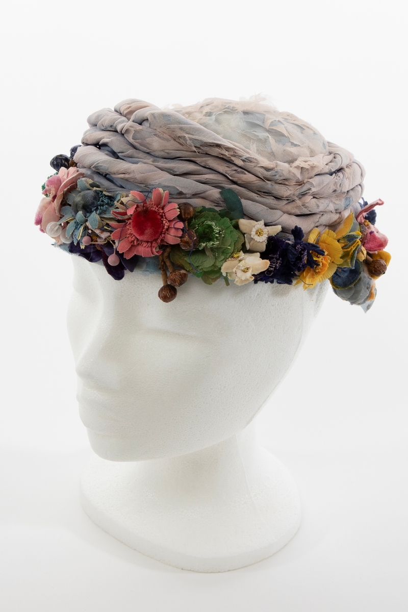 Lav, liten hatt uten brem, av lyseblå og dueblå silkechiffon. Pullen er lav og avrundet, konstruert av et grovt og mykt, hvitt nettstoff, dekket av flere lag lyseblå og tåkeblå chiffon, det ytterste laget dueblått. Mens toppen er glatt, er pullens skrå sider garnert med 5-6 snodde remser av dueblå og lyseblå chiffon. Pullens nederkant har en litt avstivet "linning", også den er trukket glatt med ciffon og nestet til resten av pullen for hånd. Nederkanten er prydet med en tett, polykrom krans av varierte blomster og blader av silke, og bær av hardt, vokset materiale. Innvendig fôret med kremfarget silke, festet for hånd, men figursydd med maskin. Svart, rund hattestrikk festet i begge sider.