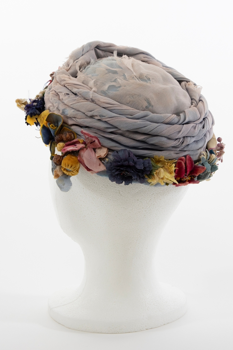 Lav, liten hatt uten brem, av lyseblå og dueblå silkechiffon. Pullen er lav og avrundet, konstruert av et grovt og mykt, hvitt nettstoff, dekket av flere lag lyseblå og tåkeblå chiffon, det ytterste laget dueblått. Mens toppen er glatt, er pullens skrå sider garnert med 5-6 snodde remser av dueblå og lyseblå chiffon. Pullens nederkant har en litt avstivet "linning", også den er trukket glatt med ciffon og nestet til resten av pullen for hånd. Nederkanten er prydet med en tett, polykrom krans av varierte blomster og blader av silke, og bær av hardt, vokset materiale. Innvendig fôret med kremfarget silke, festet for hånd, men figursydd med maskin. Svart, rund hattestrikk festet i begge sider.