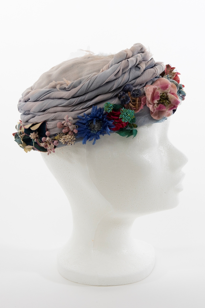 Lav, liten hatt uten brem, av lyseblå og dueblå silkechiffon. Pullen er lav og avrundet, konstruert av et grovt og mykt, hvitt nettstoff, dekket av flere lag lyseblå og tåkeblå chiffon, det ytterste laget dueblått. Mens toppen er glatt, er pullens skrå sider garnert med 5-6 snodde remser av dueblå og lyseblå chiffon. Pullens nederkant har en litt avstivet "linning", også den er trukket glatt med ciffon og nestet til resten av pullen for hånd. Nederkanten er prydet med en tett, polykrom krans av varierte blomster og blader av silke, og bær av hardt, vokset materiale. Innvendig fôret med kremfarget silke, festet for hånd, men figursydd med maskin. Svart, rund hattestrikk festet i begge sider.