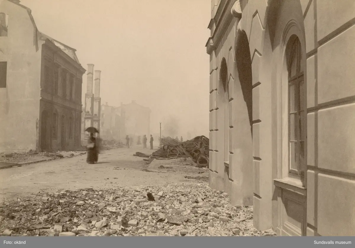 Sundsvalls stadsbrand 1888. Ensam kvinna med paraply vandrar i brandresterna efter Sjögatan med Tullpackhuset till höger.