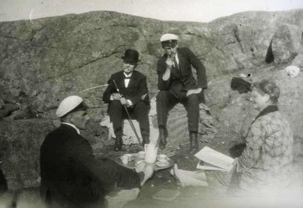 Tre män och en kvinna på picnic bland klipporna. Det står en bricka på markenmed kaffekoppar och kanna. De yngre männen röker pipa och bär studentmössor medan den äldre har käpp och kubb. Fr v:  Rolf G Bexell, advokat Otto Bexell, Gerhard Bexell och fru Gerda Bexell.