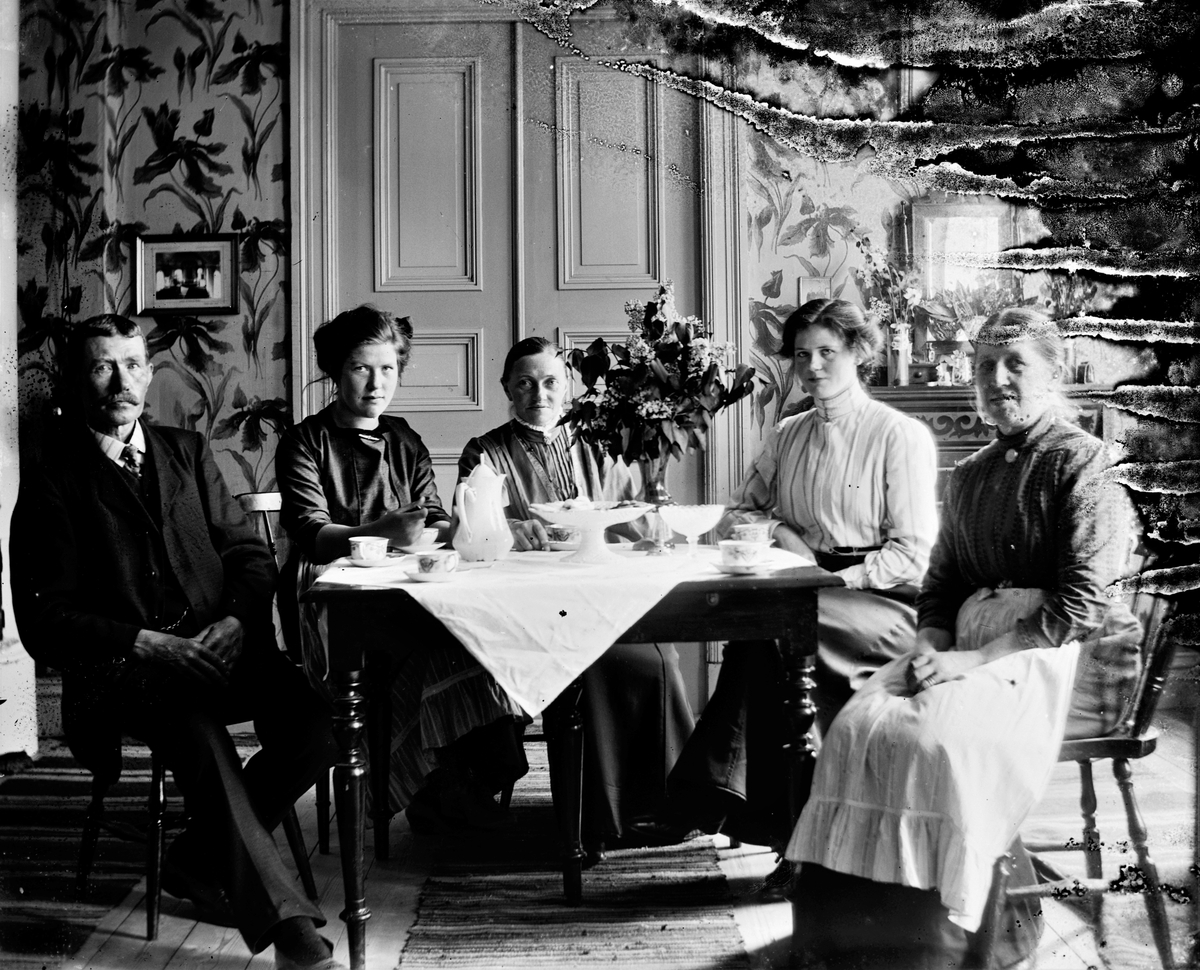 Kaffebjudning med småskollärarinnan Bertha Engström (fotografens mor) i mitten. På bordet står en stor bukett syrener, så kanske är det en skolavslutningsdag. Rummet har stormönstrade tapeter med blommor och en hög dubbeldörr (döbattang).
(Negativet skadat i högerkanten)