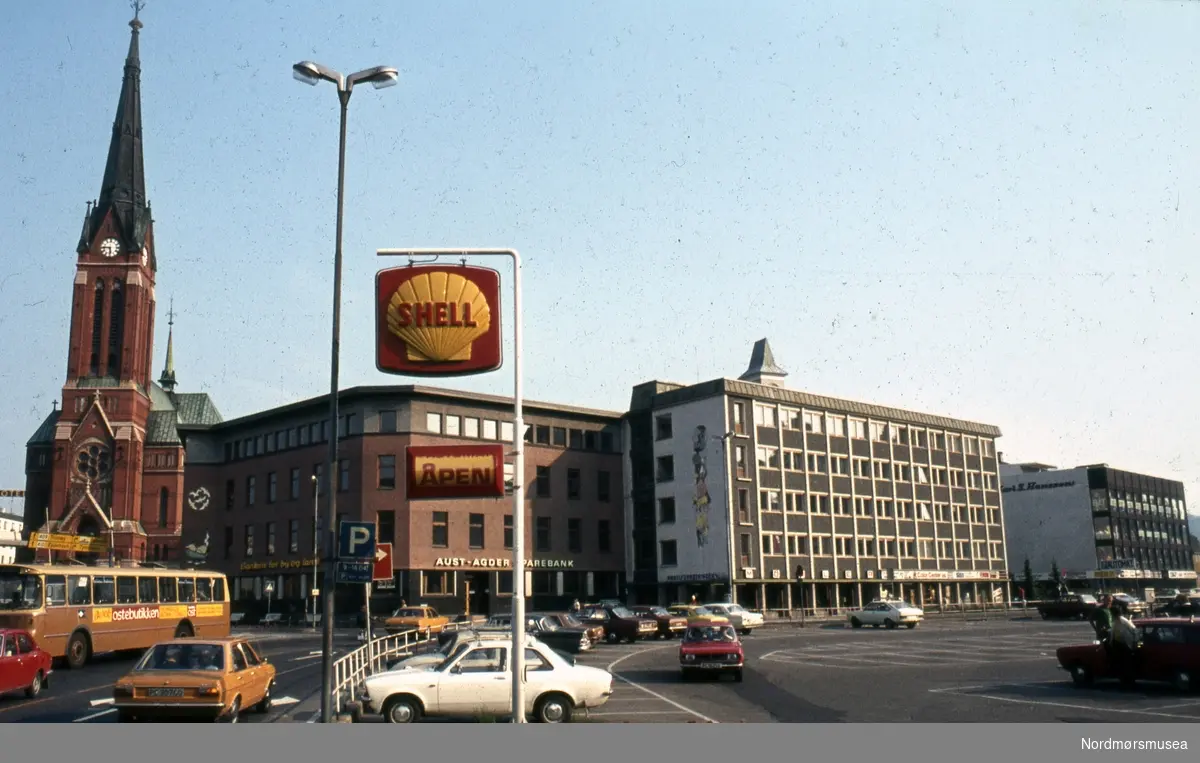 Foto trolig fra sentrum i Arendal kommune med Trefoldighetskirken og Aust-Agder Sparebank. Helt i forgrunnen et Shell-skilt.  Datering muligens omkring 1980-tallet. Fra Stein Magne Bach sin private fotosamling. Denne samlingen består av dias fra perioden omkring 1980. Fra Nordmøre museums fotosamlinger.