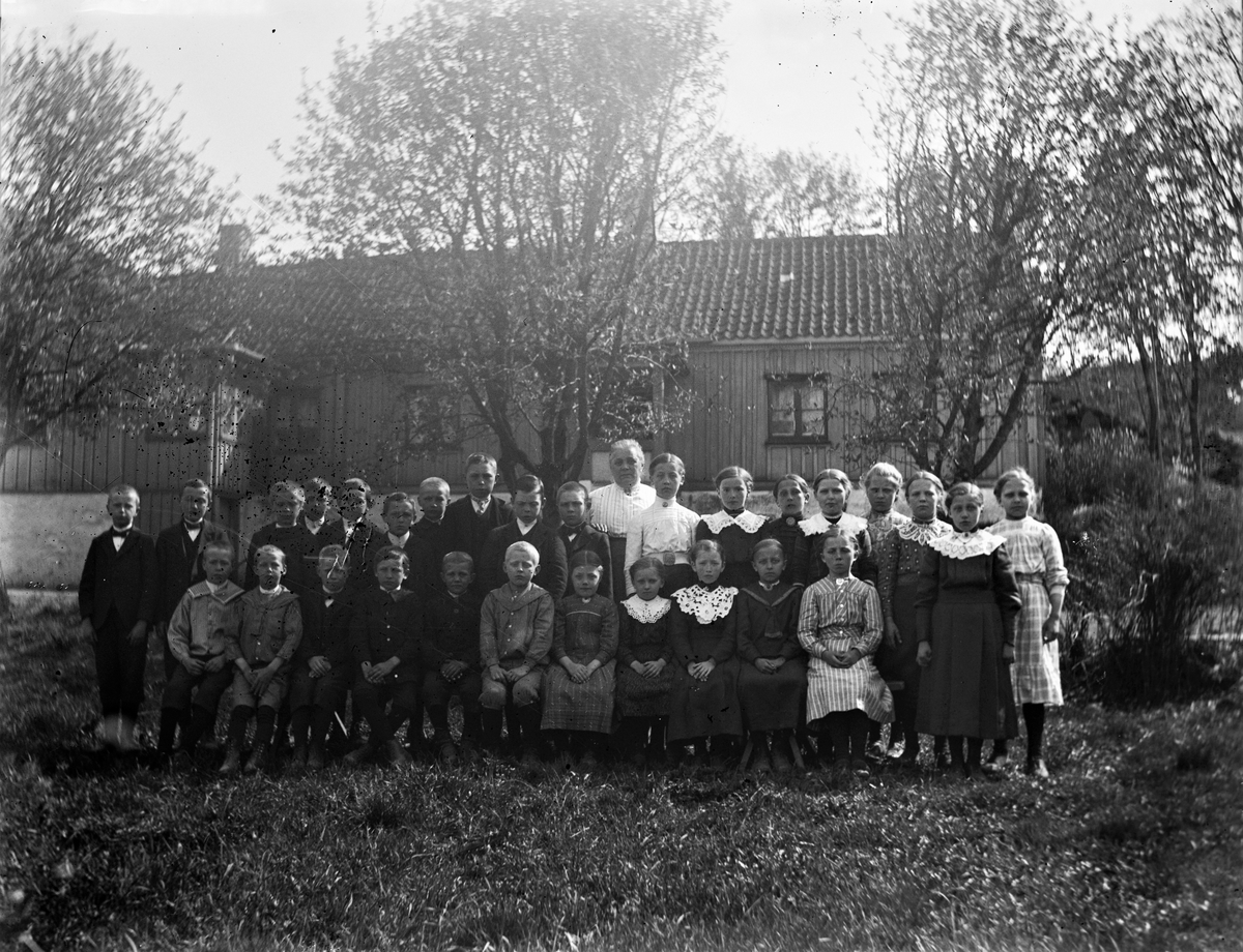 Folkskollärarinnan Constance Engström tillsammans med sina elever utanför folkskolan i Sällstorp en solig dag. Pojkarna står till vänster och flickorna till höger i bild. Längst ut till vänster står Karl-Ander Edvardsson och som nr 4 till höger om Constance står flickan som skulle komma att gifta sig med honom: Alma Olivia Gustavsdotter.
(Negativet skadat till vänster)