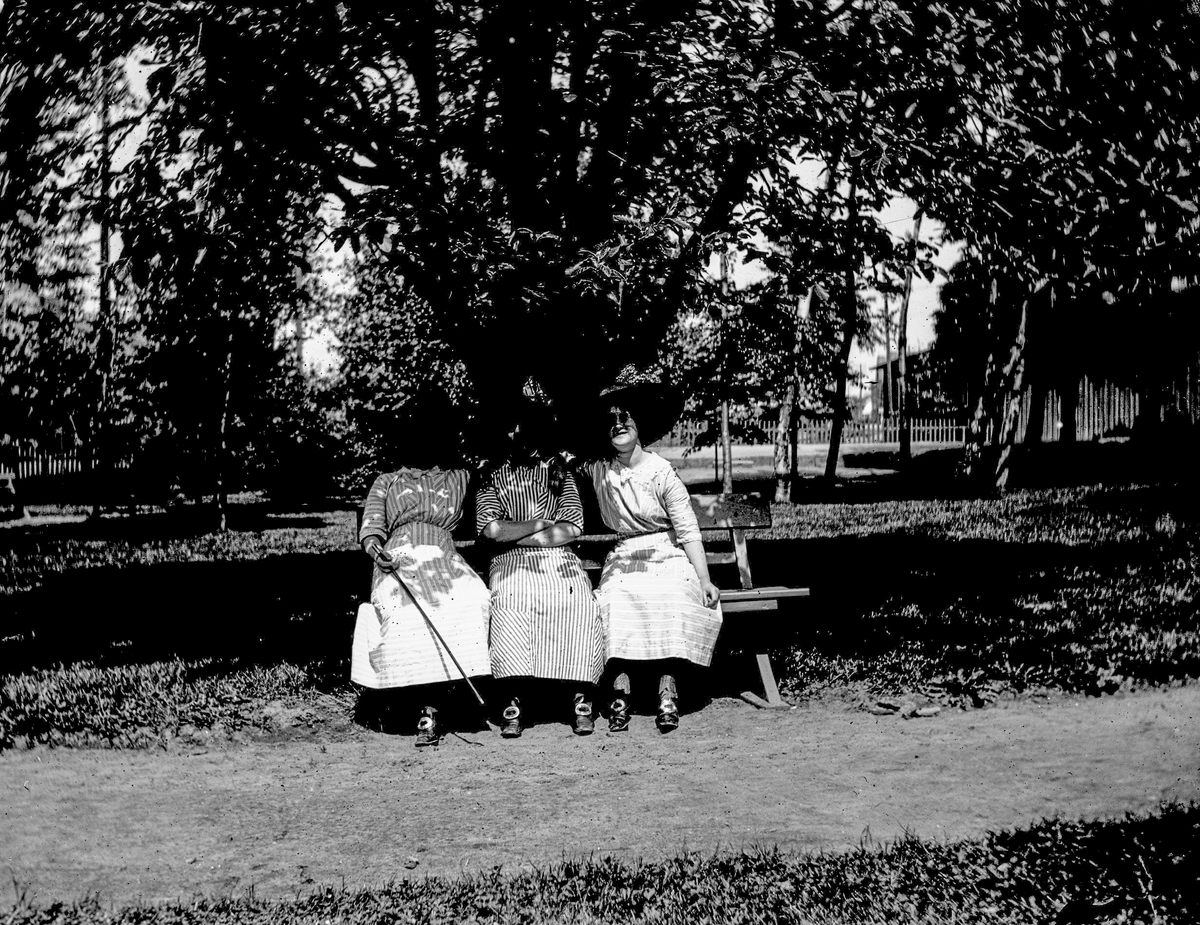 Tre kvinnor sitter på en parkbänk, kanske i Varbergs Societetspark. Skuggan är så djup att endast ansiktet på kvinnan längst till höger går att skönja. De yttre vilar kamratligt sina händer på sin vän i mitten. Alla tre bär likadana skor med stora spännen och klänningar i olika randade tyger.