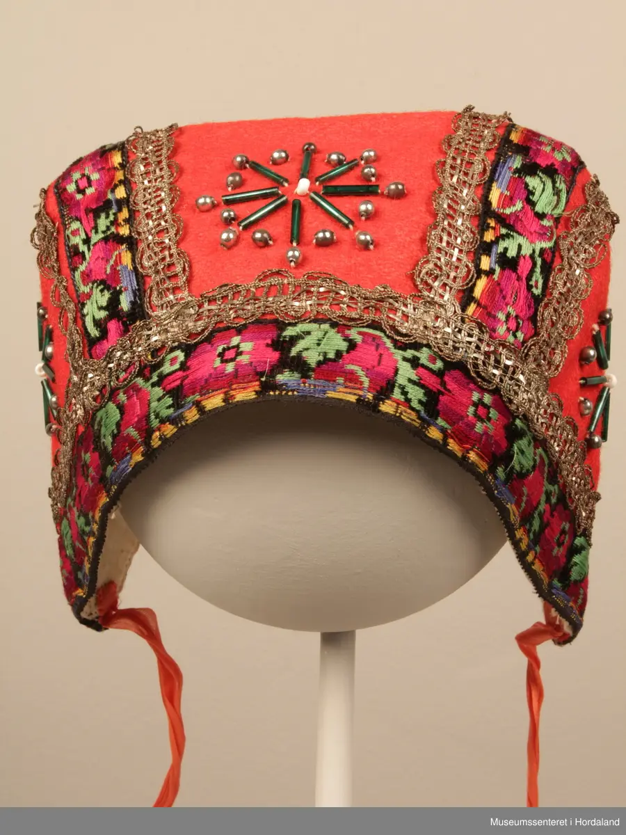 Raud barnebunadshue med påsydde perler, roseband og sølvfarga metallband. Stjernemotiv av grøne stråperler. Store sølvarga, runde perler rundt desse, kvit i midten. Roseband til kanting og på dei to saumane som går bakover. Metallbord i bølgjer på kvar side og ovanfor kantbandet. Kvitt bomullsfôr. Raude knyteband i kvar side.