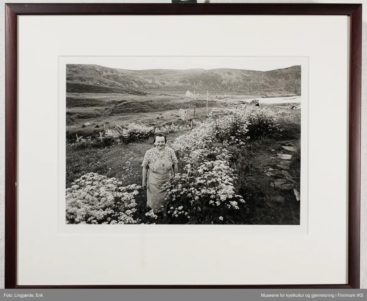 En dame står blant blomster. I bakgrunnen ser man litt bebyggelse og små båter liggende i fjære.