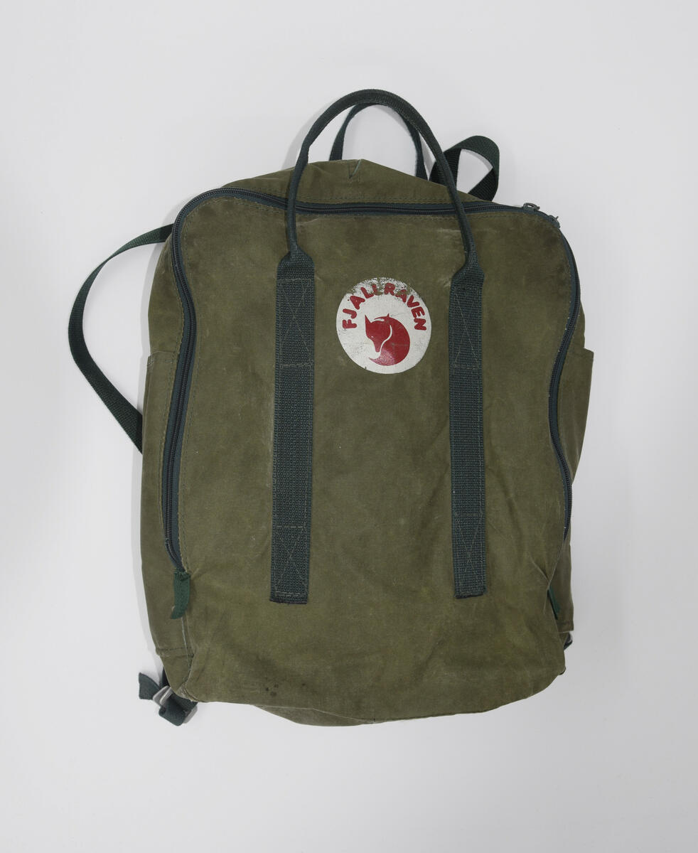 Ryggsäck i modellen Kånken. Grön. Fyrkantig i formen mett ett fack med dragkedja längst långsidorna och upptill. Ett mindre fack inne i det stora facket.Två par handtag på bak och framsida som går över ovansidan. Justerbara axelremmar, två stycken, av tuskaft vävt band. 
Märkte, rund, på framsidan med texten Fjällräven och företagets logga i form av en Fjällräv. 
Dragkedja och övriga detaljer av metall.