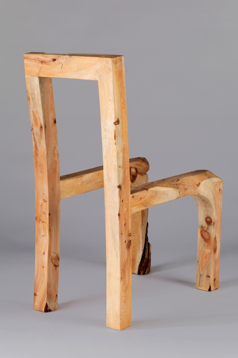 Skulpturelt objekt i tre, utformet som en stol uten sete. Høyreist rygg som består av to stolper som glir over i bakbeina, og forbundet hverandre med et toppstykke. Sarg og forbein er laget av samme treemne.