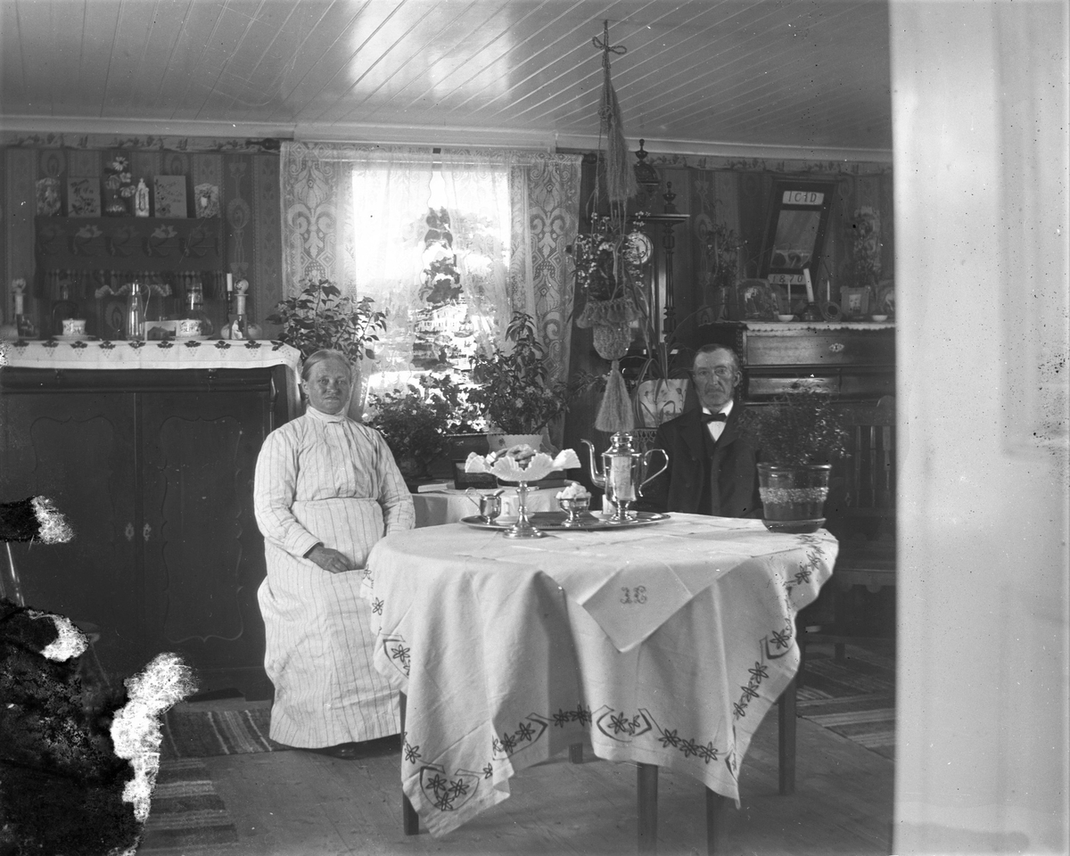 Ett äldre par sitter vid bordet i ett ombonat finrum som har två skänkar med prydnadssaker såsom fotografier och karaffer, en spegel med initialerna ICID och årtalet 1870, väggur och en textilampel som hänger från taket. Bordet har dubbla broderade dukar och på brickan står en kaffeservis i metall bredvid kakfatet på hög fot med veckad glasskål. Både tapeter och textilier bär jugendprägel men tapetbårdens dekor avviker med motiv av väderkvarnar och segelbåtar. I fönstret är rullgardinen med en hus- och landskapsdekor nerdragen.
(Möjligen samma par på bildnr GEB238)