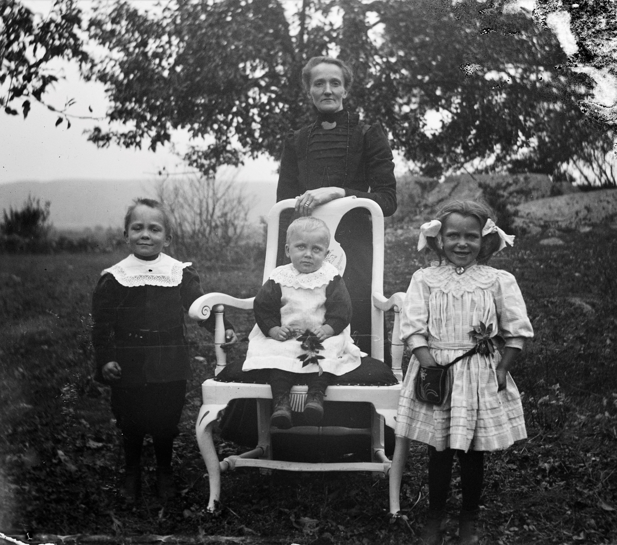 Anna Petronella Johansson (född Andersdotter) med tre av sina sju barn samlade utomhus, Stora Råryd. Minsta barnet Erik August sitter med en kvist i knät på en länsstol i bondrokoko med tvillingarna Hulda Andrea och Karl Ragnar på var sin sida. Alla tre har spetskragar av olika slag, dottern i somrig klänning, hårrosetter, blomma vid skärpet och en liten väska med blomsterbrodyr på ena sidan och hennes tvillingbror i sammetsaktig mörk jacka med brett skärp och puffärmar. (Se även bildnr GEA119)