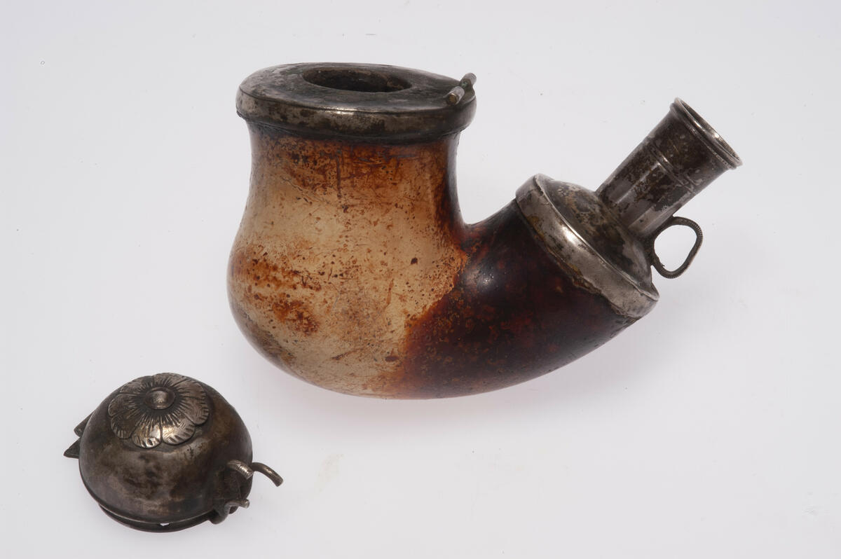 Piphuvud, av sjöskum. Silverbeslag runt skafthål och rökhål, samt kupigt lock med påfäst 8-uddig blomma. Stämplar: ESH H C5 = Eric Söderholm Härnösand 1857. Gåva: Fru häradshövding Grufman, Stockholm 1960, (samling tillhört fadern distriktslantmätare A.H.L. Andersson, Sollefteå). 