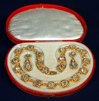 Krans av guld med dekorationer föreställande små blommor i form av fyra runda rubiner samt fyra blommor med blåmålade blad. I mitten en rund månsten. Smycket sätts på örat med hjälp av en guldpinne vilken man sticker genom ett hål i örsnibben och fäster fast med en liten knopp som sätts på pinnens ände. Från kransen hänger i en länk en likadan men ovalformad krans och med en större ovalformad i mitten. Tillhör halsband inv.nr 34617.