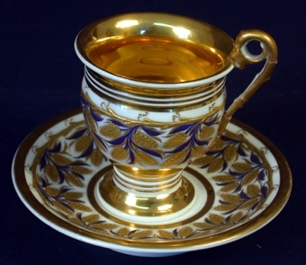 Material 1: Porslin. Material 2: Guld.
:1 Kopp av vitt porslin med stora ytor täckta med förgyllning i form av dekorativa bårder samt runt hela koppen ett lövverk med blad i guld och blå målad färg. Större delen av koppens insida är förgylld. Ett snirkligt öra med vilket man håller i koppen när man dricker är med ena änden fäst på öppningens kant och den andra änden fäst längre ner på koppens sida. Koppen står på en 20 mm hög fot, diameter: 53 mm. Koppens hela höjd: 86 mm, djup: 75 mm. Öppningens kant något utåtböjd. Diameter i öppning: 68 mm, i botten: 57 mm. 
 :2 Kaffefat i vitt porslin, diameter: 144 mm. Fatets ovansida täckt med ett lövverk med blad i i guld och blå målad färg samt ett guldband runt kanten. 
    Tillhör servis där inv.nr. 34621-34625 ingår.