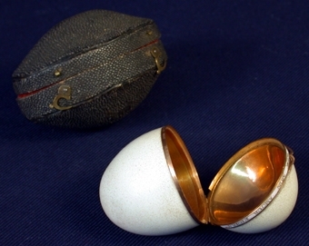Material 1: Äggskal.
Material 2: Förgylld metall.
Material 4: Textil: Sammet.
 :1 Ask i form av ett tomt äggskal från okänd fågel, delat på mitten till en bottendel med lock. Hela äggskalets insida täckt med förgylld metall tillika de två delarnas kanter. Locket sitter fast på underdelen och kan öppnas och stängas med hjälp av ett litet gångjärn. Runt hela ägget ligger på utsidan ett band av förgylld metall vars mitt är vit. På den vita ytan står med guldversaler: "Quand il eclora mon amour fintra". Mellan bokstäverna finns bladliknande mönster. Äggets omkrets: 145 mm.
   :2 Ask att förvara ägget i. Äggformat av brunt plastliknande konstmaterial med ojämn yta. Bottendel med lock som öppnas och stängs med hjälp av ett gångjärn. På bottendelens utsida två krokar att trä in i märlor på locket, vid låsning av asken. Insidan klädd med rött sammetstyg. Höjd: 55 mm, bredd: 51 mm, längd: 80 mm, omkrets: 170 mm.