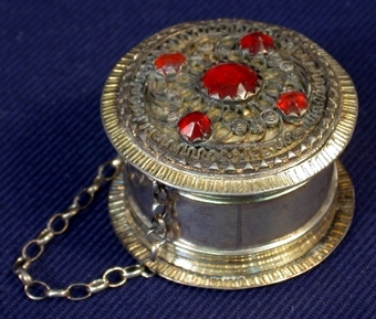 Cylinderformad, av metall ståendes på hög kant med platt botten. Ovansidan dekorerad med tennbroderi och fem pärlor av rubin. En tunn metallkedja är fäst med ena änden i ovansidans kant och den andra i botten. På undersidan är graverat med versaler: "KKS" och "BSD" och i metallen är stämplat: "CEL N G" samt två märken.