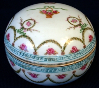 :1-2 Rundformad skål där lock och underdel ser likadana ut. Av vitt porslin. På utsidan dekorerad med målade mönster i blått, samt rosa rosor, gröna blad, rosetter, kransar m.m. På skålens undersida finns märket för den österrikiska porslinfabriken Vienna stämplat, en bikupa i blått.