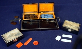Material 4: Papper.
:1 Bricka i trä på vilken fyra askar med spelmarker i förvaras. Handtag av metall. På brickans insida två lister i kors som bildar fack på brickan att placera askarna i. Längd: 220 mm, bredd: 127 mm, höjd: 20 mm.
   :2 Ask av trä med lock som kan öppnas och stängas med hjälp av gångjärn i metall. Stängs igen med hjälp av beslag på framsidan. Lockets ovansida består av ett fönster i plast med metallram runtom. Innanför fönstret en teckning i blyerts på papper, föreställande ett bergslandskap med vatten, båt och hus. Bredd: 80 mm, djup: 52, höjd: 26 mm. 
   :3-18 16 stycken tunna rektangulära spelmarker i vitt benmaterial (elfenben?), i två storlekar; 13 stycken: längd: 44 mm, bredd: 13 mm, 3 stycken: längd: 26 mm, bredd: 18 mm. Förvaras i ask :2.
   :19 Ask likadan som :2 men teckningen på lockets ovansida föreställer en bro över en å med frodigt buskage runtom samt en kupolliknande byggnad i bakgrunden.
   :20-44 25 stycken tunna spelmarker i rödfärgat benmaterial (elfenben?), i tre storlekar och två former; 15 stycken rektangulära: längd: 44 mm, bredd: 13 mm, 5 stycken rektangulära: längd: 26 mm, bredd: 18 mm, 5 stycken cirkelrunda, diameter: 22 mm. Förvaras i ask :19.
   :45 Ask likadan som :2 men teckningen på lockets ovansida föreställer en klippformation i en sjö med frodigt buskage runtom. 
   :48-70 25 stycken tunna spelmarker i mörkbrunfärgat benmaterial (elfenben?), i tre storlekar och två former; 15 stycken rektangulära: längd: 44 mm, bredd: 13 mm, 5 stycken rektangulära: längd: 26 mm, bredd: 18 mm, 5 stycken cirkelrunda: diameter: 22 mm. Förvaras i ask :45.
   :71 Ask likadan som :2 men teckningen i på lockets ovansida föreställer två hyddliknande byggnader med halmtak, med träd och buskar runtom.
   :72-91 20 stycken tunna spelmarker i blåfärgat benmaterial (elfenben?), i tre storlekar och två former; 16 stycken rektangulära: längd: 44 mm, bredd: 13 mm, 3 stycken rektangulära: längd: 26 mm, bredd: 18 mm, 1 stycken cirkelrunda. Förvaras i ask :71.